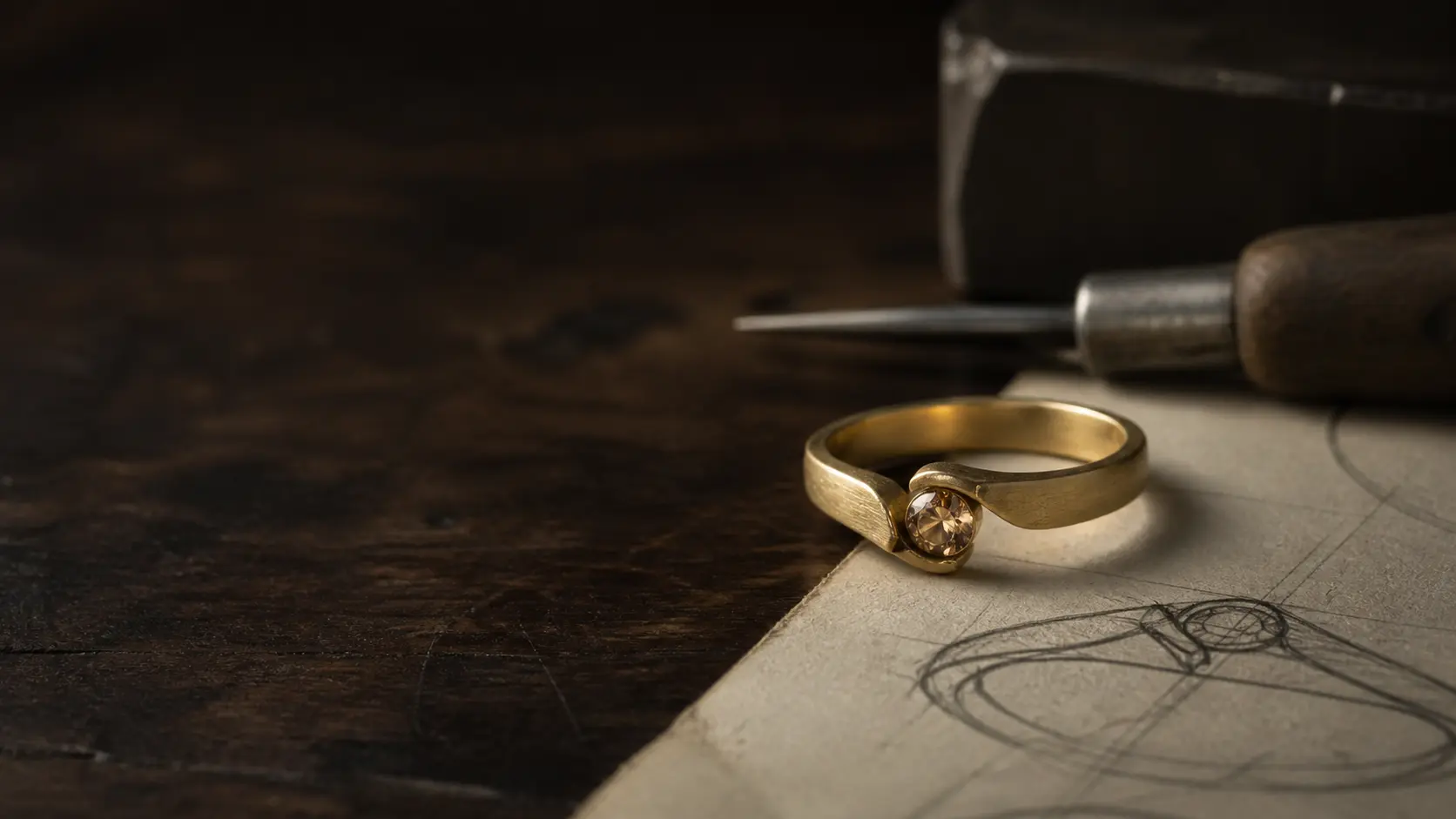 Gold ring resting on a hand-drawn jewelry sketch at a worn jeweler's bench
